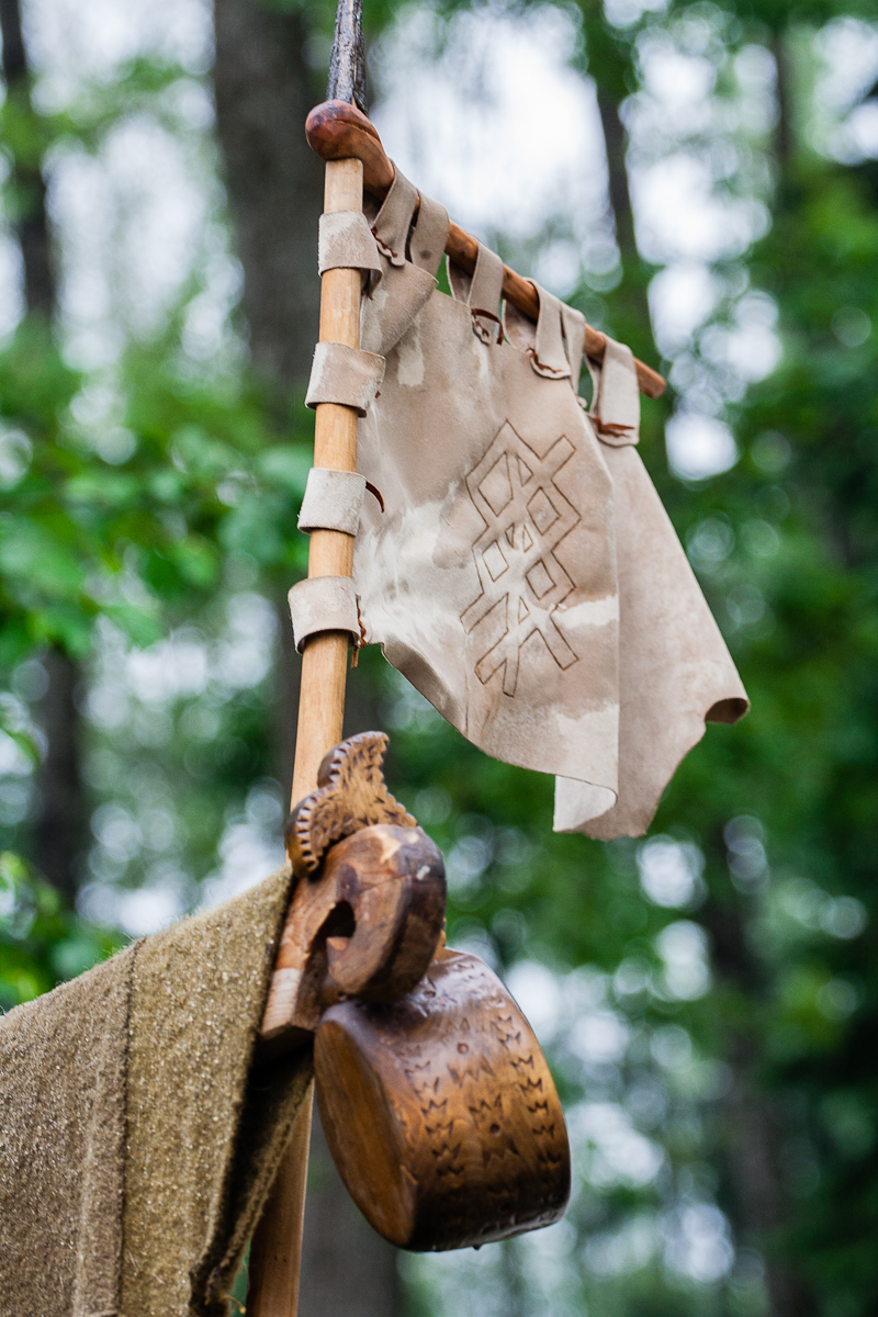 A tattered, ancient-looking beige flag with a geometric design hangs from a wooden pole adorned with leather straps. Below the flag, there is a wooden drinking vessel with intricate carvings. Set against the backdrop of a dense green forest, this scene evokes memories of Kilkim Žaibu 2012 festival.