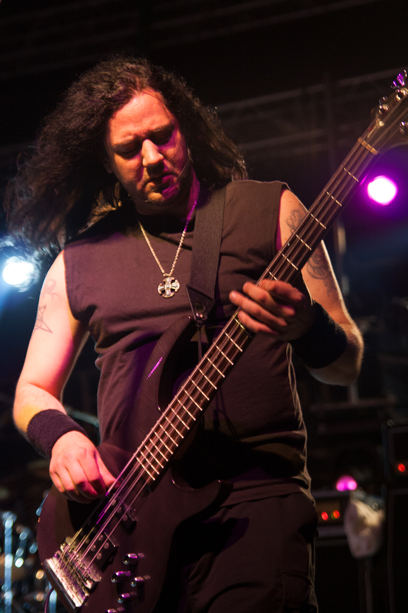 A musician with long dark hair is playing an electric bass guitar on stage at the 2012 Kilkim Žaibu festival. He is wearing a sleeveless black shirt, black wristbands, and a necklace with a pendant. The stage is lit with colorful lights, including purple and blue, creating a vibrant atmosphere.