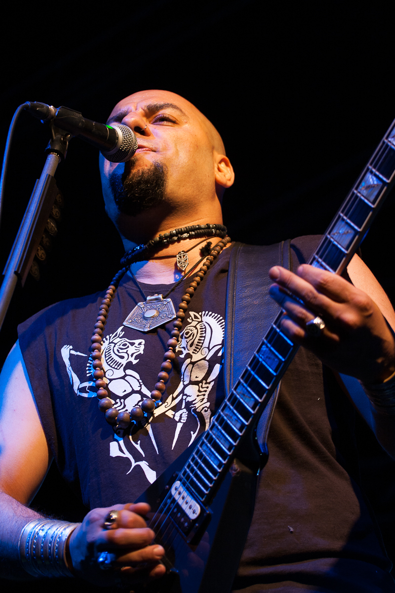 A bald musician with a goatee is playing an electric guitar and singing into a microphone at the 2012 Kilkim Žaibu festival. He is wearing a sleeveless black shirt with a graphic design and layered necklaces, including beads and an amulet. The dark background highlights him with stage lighting.