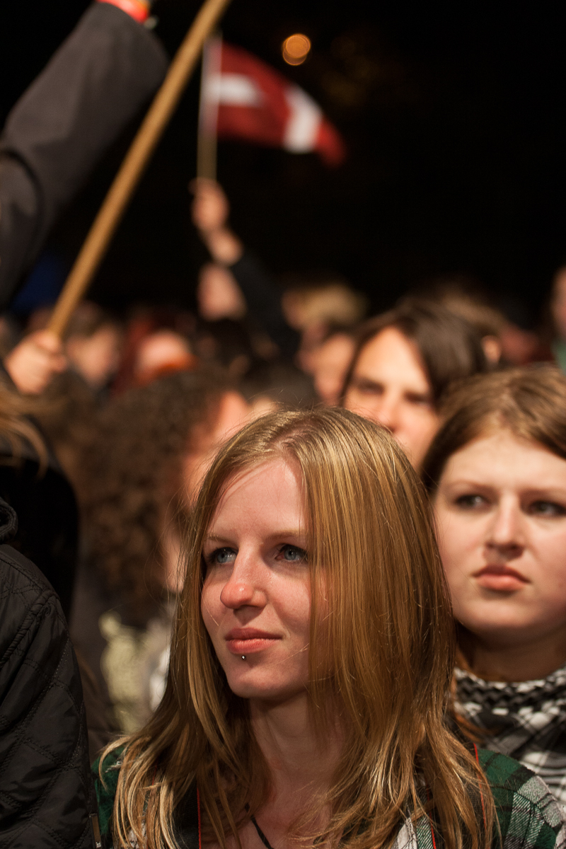 At the 2012 Kilkim Žaibu festival, a blonde woman in a green jacket stands among a dense crowd, looking ahead with a thoughtful expression. Another young woman behind her has long dark hair and a neutral look. The crowd appears focused, and in the background, someone holds up a red and white flag.
