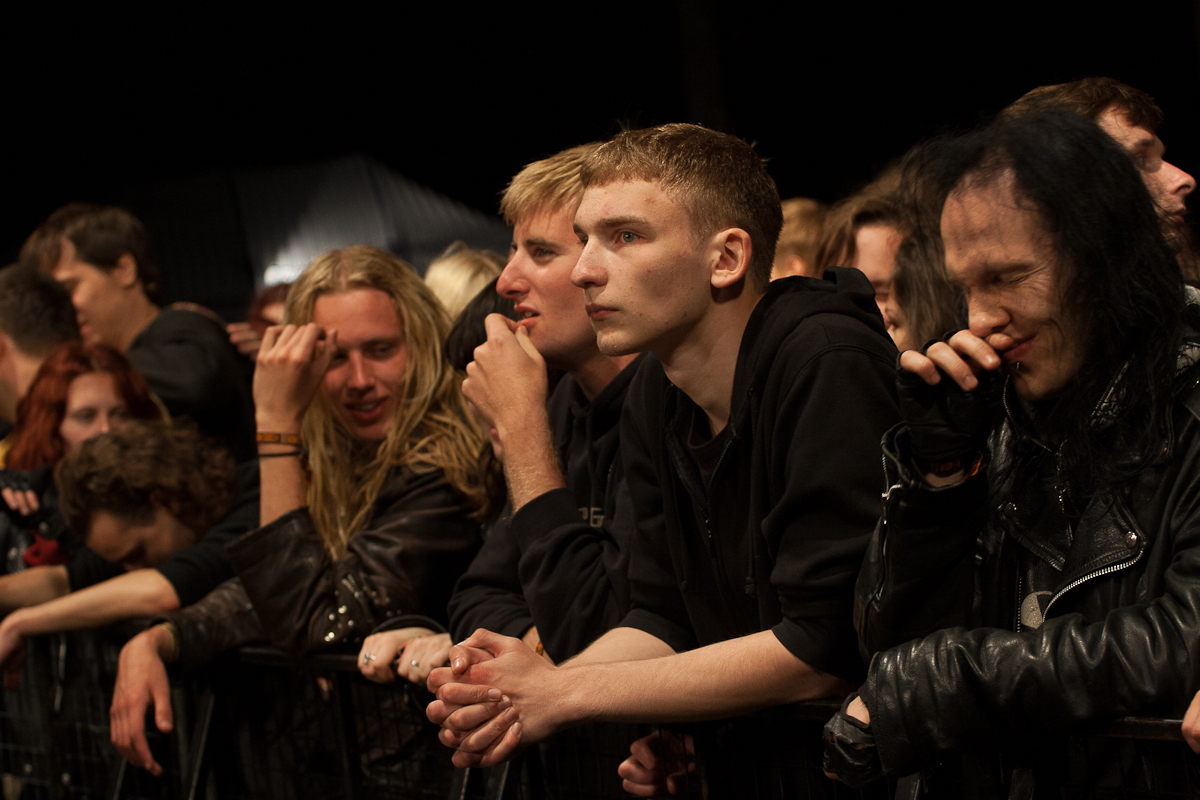 A group of concertgoers, predominantly young adults, is closely packed together behind a barricade at an outdoor nighttime event. They are attentively watching the stage, dressed in dark casual attire, some with long hair and others with short hair. The atmosphere at the 2012 Kilkim Žaibu festival suggests anticipation and excitement.