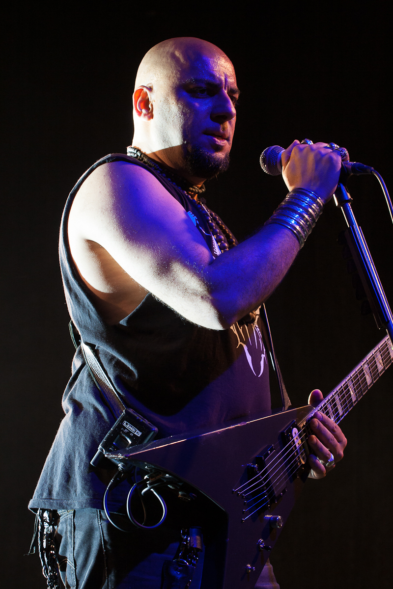 A bald musician with a goatee passionately sings into a microphone while playing a V-shaped electric guitar at Kilkim Žaibu 2012. He wears a sleeveless black shirt, and his wrists are adorned with multiple metal bracelets. The festival stage lighting highlights his arms and face against a dark background.