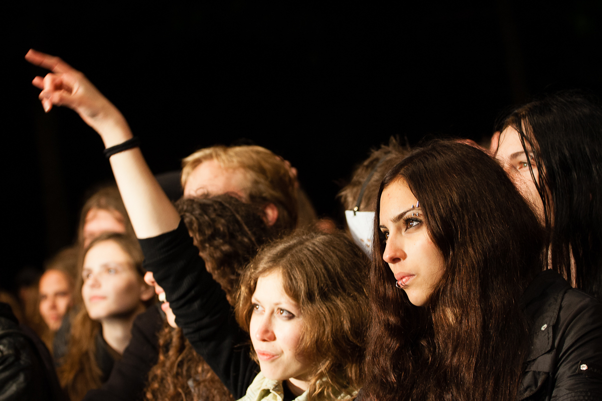 A crowd of people at a festival, mostly young adults, some with long hair. A woman in the foreground has dark, straight hair and a serious expression, while another woman nearby, with curly hair, raises her arm excitedly. The background is dark, highlighting the individuals' faces with Kilkim Žaibu 2012 concert lighting.
