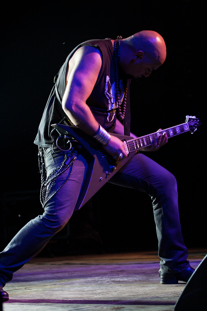 A bald musician wearing a sleeveless black shirt, jeans, and boots is passionately playing an angular electric guitar on stage at Kilkim Žaibu 2012. He is leaning forward with one leg bent, creating an intense and dynamic pose. Chains and wristbands add to his rock or heavy metal aesthetic. The background is dark.