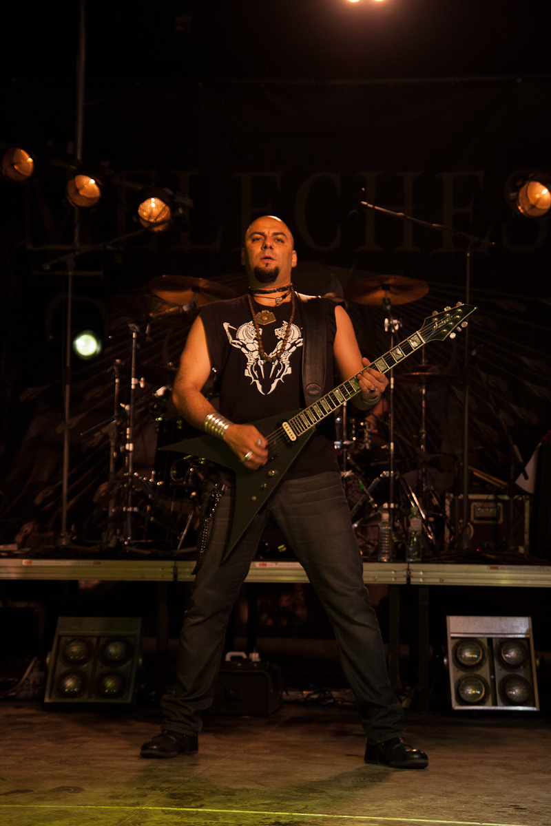A musician in a black sleeveless shirt with a white graphic plays a black, V-shaped electric guitar on a dimly lit stage at the 2012 Kilkim Žaibu festival. He wears dark pants and stands in front of a drum set. Overhead stage lights illuminate him as he performs, with part of the band banner in the background.