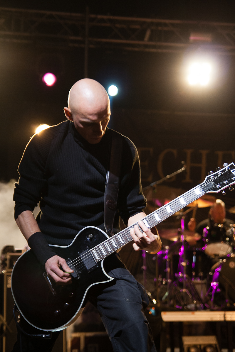 A bald musician wearing a black shirt and wristband is intensely playing an electric guitar on stage under bright lights at Kilkim Žaibu 2012. A drum set and another band member are visible in the background. The stage is illuminated with colorful lights, creating a vibrant atmosphere.