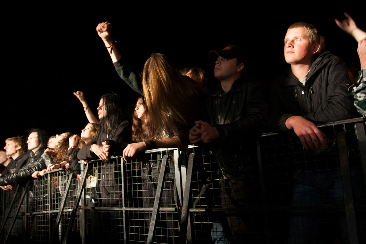 Concertgoers stand behind a metal barricade at an outdoor nighttime event in Lithuania. Several have their arms raised, some with clenched fists. Dressed in dark clothing, the lighting captures their enthusiastic expressions as they engage with the performance, reminiscent of Kilkim Žaibu 2012 festival vibes.