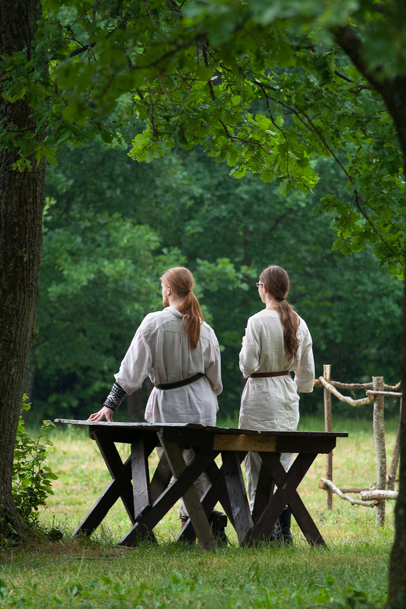 Two people with long hair stand near a wooden bench in a forest clearing. Dressed in medieval-style clothing reminiscent of Kilkim Žaibu 2012, they face away from the camera, towards a sunlit, green background. A tree's branches frame the top of the image, providing partial shade. Wooden structures are visible in the distance.