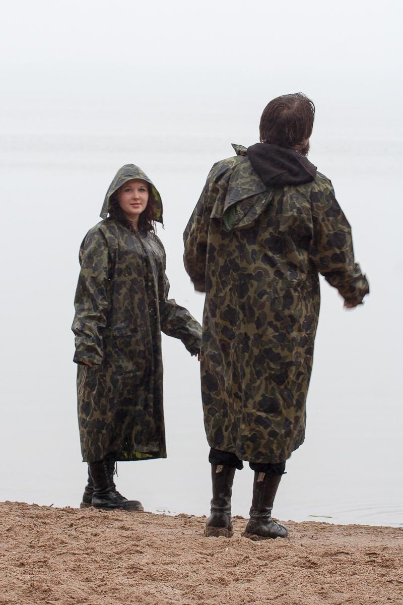 Two people wearing long camouflage raincoats and black boots stand on a sandy beach during Kilkim Žaibu 2012. One person faces the camera with their hood up, while the other has their back turned, looking toward the water. The overcast sky suggests a rainy or misty day.