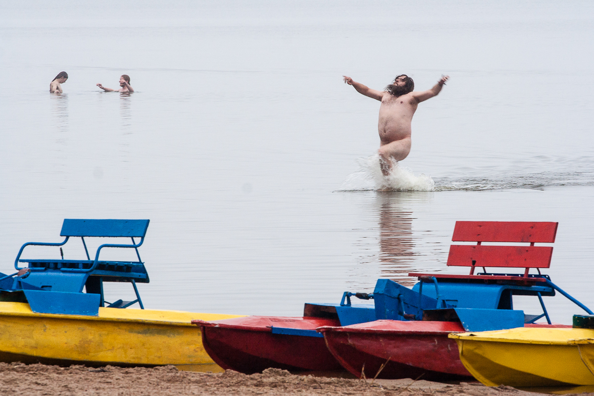 A man joyfully runs out of the water with arms raised at a beach, clearly enjoying the moment. Two other people are seen further in the water, engaged in conversation. In the foreground, brightly painted red, yellow, and blue paddle boats are lined up on the sandy shore—a perfect scene from Kilkim Žaibu 2012.