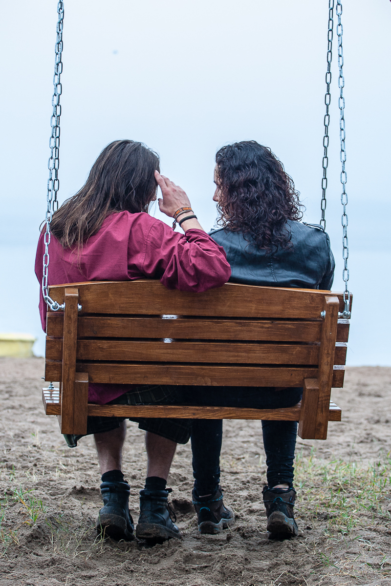 Two people with long hair sit side by side on a wooden swing overlooking a body of water, reminiscent of a serene moment captured at Kilkim Žaibu 2012. The person on the left wears a red jacket and black boots, while the one on the right sports a black leather jacket. They sit closely together, with the person on the left raising their hand to their head.