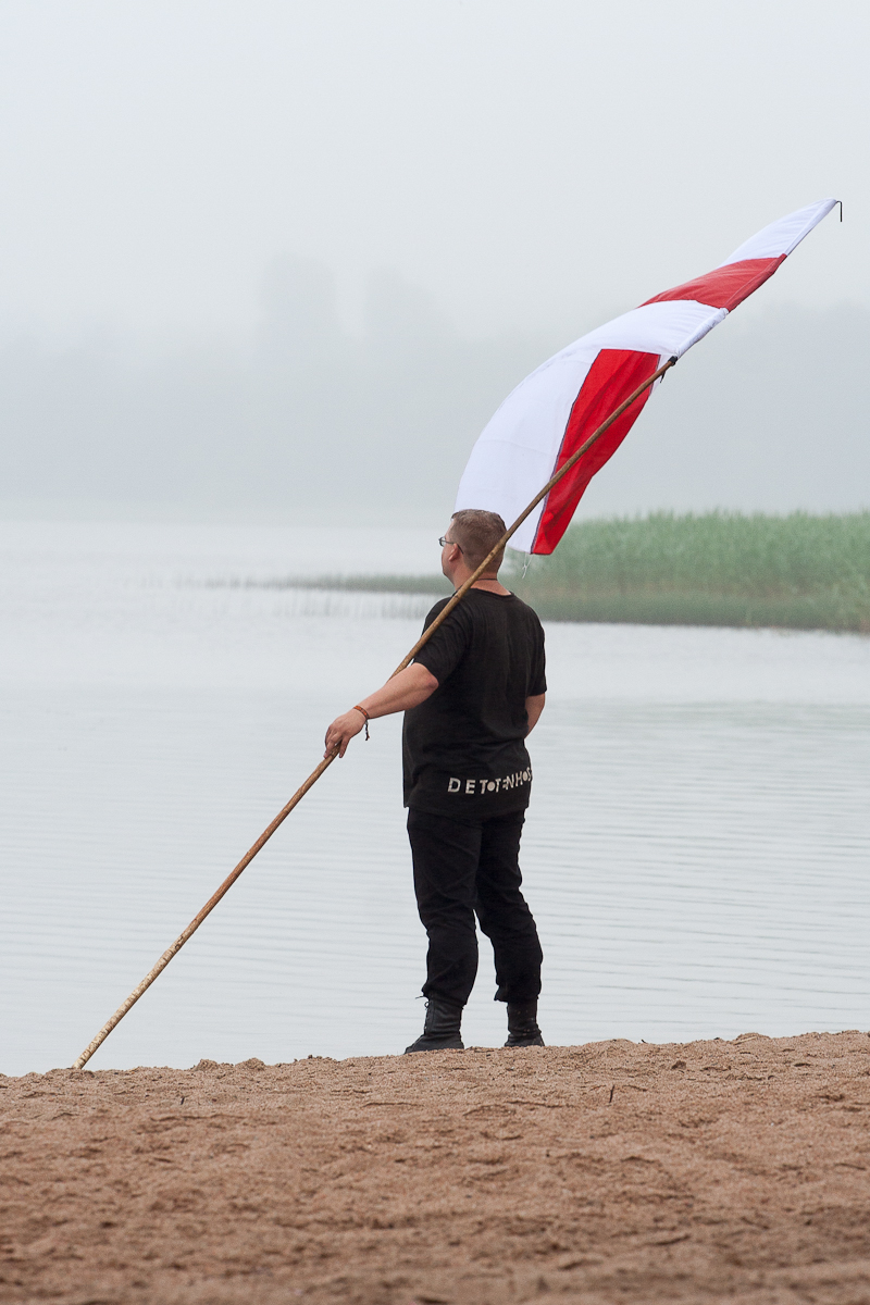 A person standing on a sandy beach holds a large red and white flag mounted on a long pole. The individual, dressed in black with "DETOX" written on the back of their shirt, seems to be part of the 2012 Kilkim Žaibu festival. The background features a foggy lake with blurred vegetation in the distance.