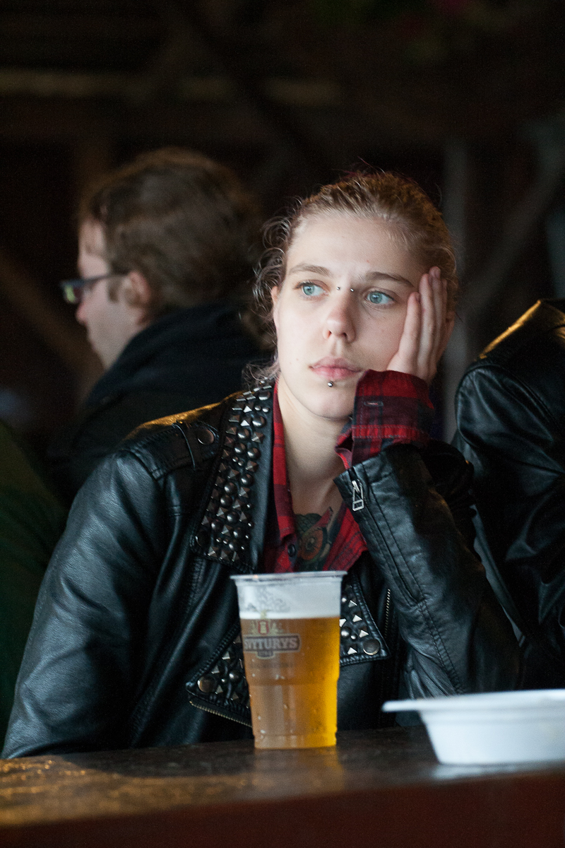 A young person with light brown hair, wearing a studded black leather jacket and a red plaid shirt, rests their head on their hand. They are seated at a table with a plastic cup of beer in front of them at Kilkim Žaibu 2012 festival. Their expression appears contemplative while another person is visible in the background.