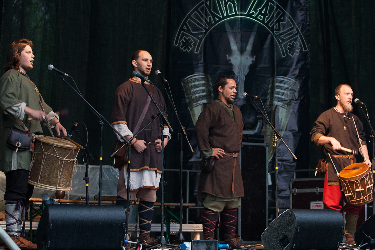 Four men dressed in medieval-style clothing stand on a stage at the 2012 Kilkim Žaibu festival. Two of them play drums while the others sing into microphones. Behind them is a banner featuring a goat skull and the word "Vikivarri" in stylized letters. The dark backdrop emphasizes the performers.