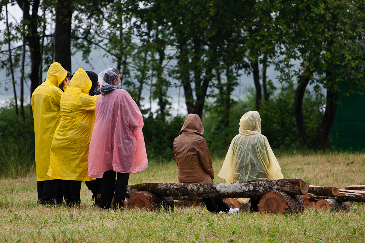 A group of people wearing yellow, pink, and clear rain ponchos stands and sits in a grassy area near logs at Kilkim Žaibu 2012. The ponchos shield them from the rain as they enjoy the misty, damp outdoor setting. Some festival-goers are seated on the logs, while others stand nearby.
