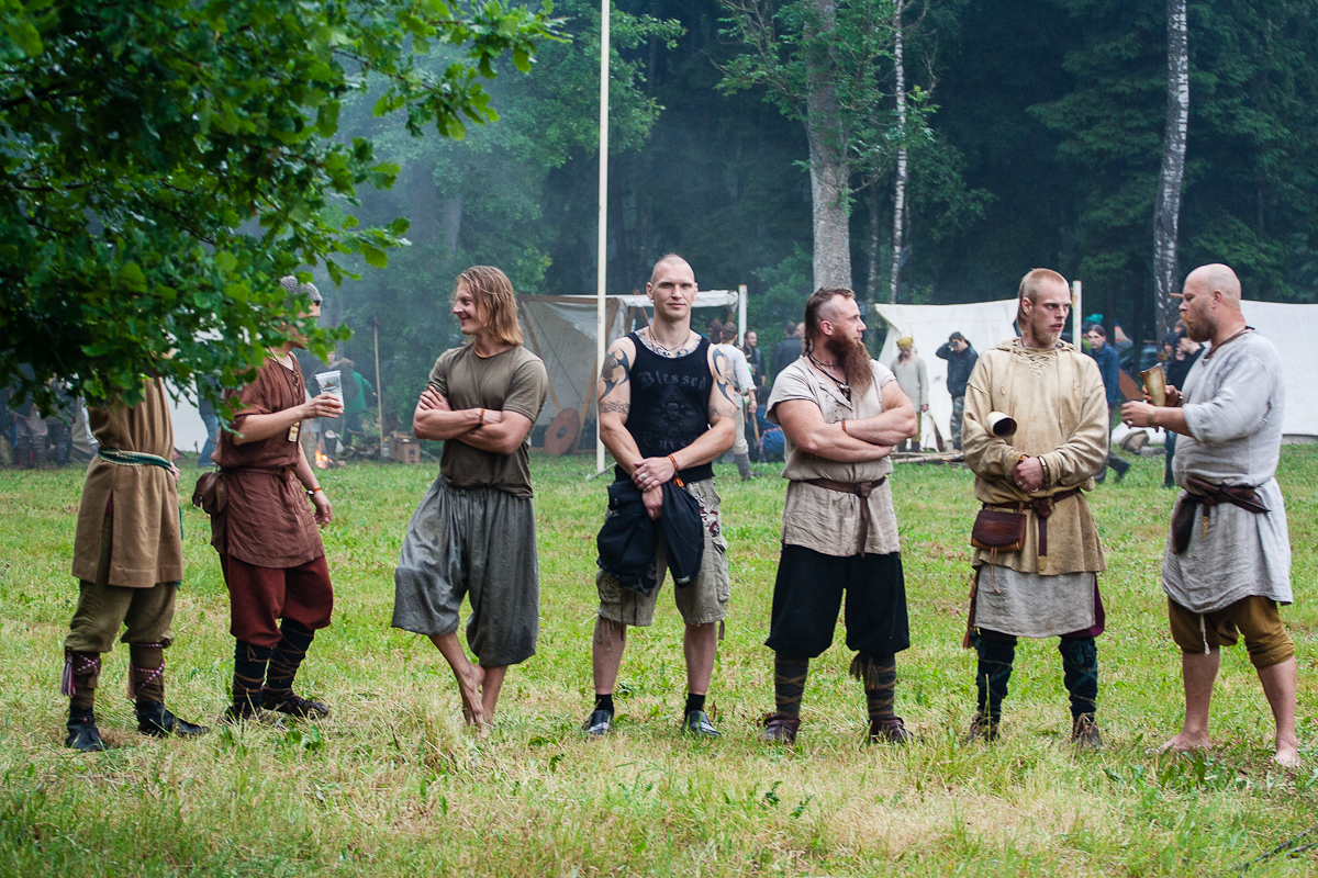 A group of six men dressed in medieval-style clothing stands outdoors on a grassy field with trees and tents in the background at Kilkim Žaibu 2012. They are engaged in conversation and holding various objects. Some have tattoos, and one has a long, braided beard. Their outfits include tunics, belts, and boots.