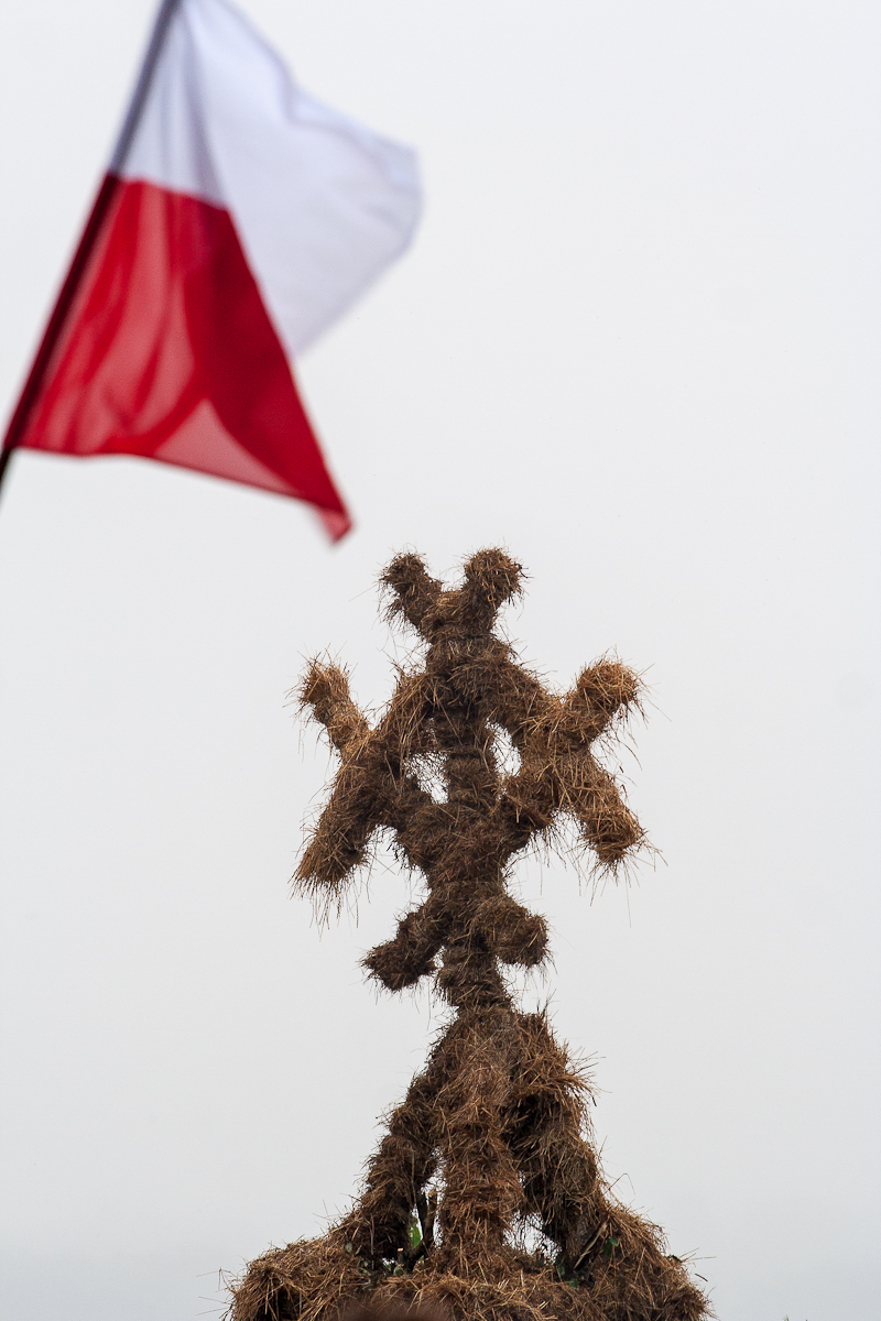 A structure made of hay bales is arranged in a cross-like pattern against a white sky background. In the upper left corner, a red and white flag, reminiscent of the 2012 Kilkim Žaibu festival, is partially visible, fluttering in the wind. The image has a minimalistic and rustic atmosphere.