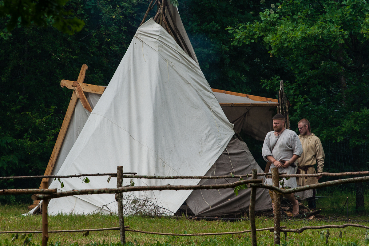 Two men dressed in medieval-style tunics stand outside a large white tent set up in a grassy area with dense green trees in the background. Propped up with wooden poles, the tent and rustic wooden fence create a scene reminiscent of Kilkim Žaibu 2012, adding authenticity to the historical reenactment.