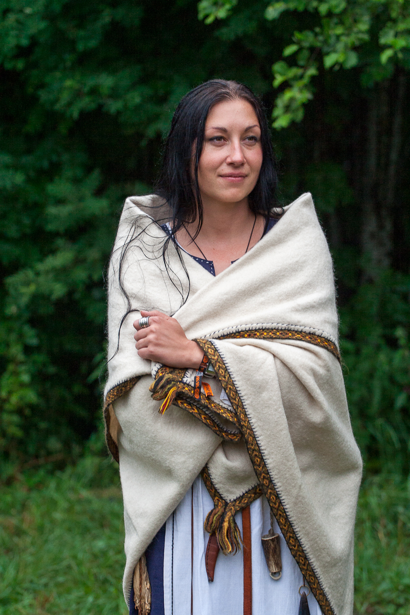 A woman with long black hair stands in front of a lush, green forest background at the 2012 Kilkim Žaibu festival. She is wrapped in a cream-colored blanket adorned with decorative trim. Gazing slightly to the left with a calm expression, her clothing underneath the blanket is white with dark accents.