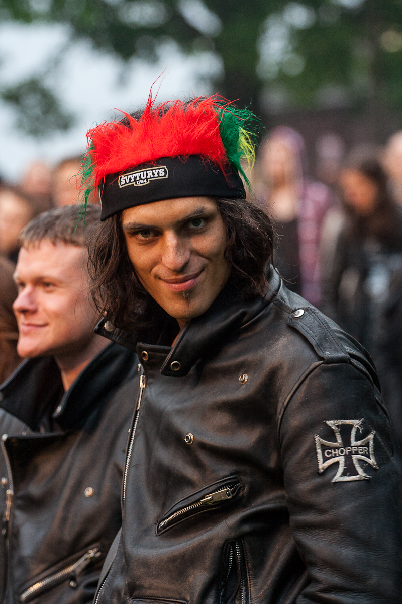 A person with long hair, wearing a headband adorned with red and green feathers and a 5V TURYS label, smiles at the camera at the 2012 Kilkim Žaibu festival. They are dressed in a black leather jacket with a "Chopper" patch on the sleeve. Another person in similar attire is partially visible in the background.