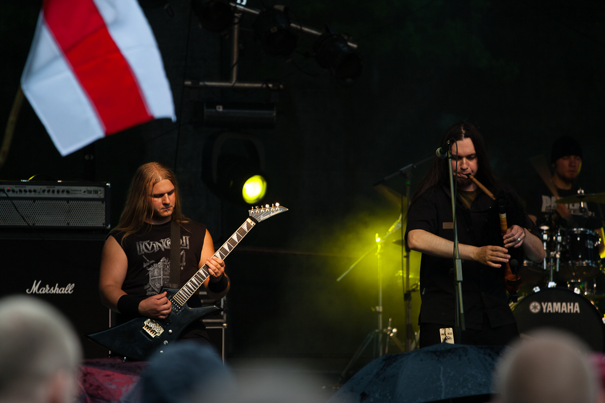 Three musicians perform on an outdoor stage at Kilkim Žaibu 2012. The guitarist on the left wears a black sleeveless shirt and plays an electric guitar. The center musician, clad in dark clothing, passionately plays bagpipes. The drummer in the background is partially obscured as a white and red flag waves in the foreground.
