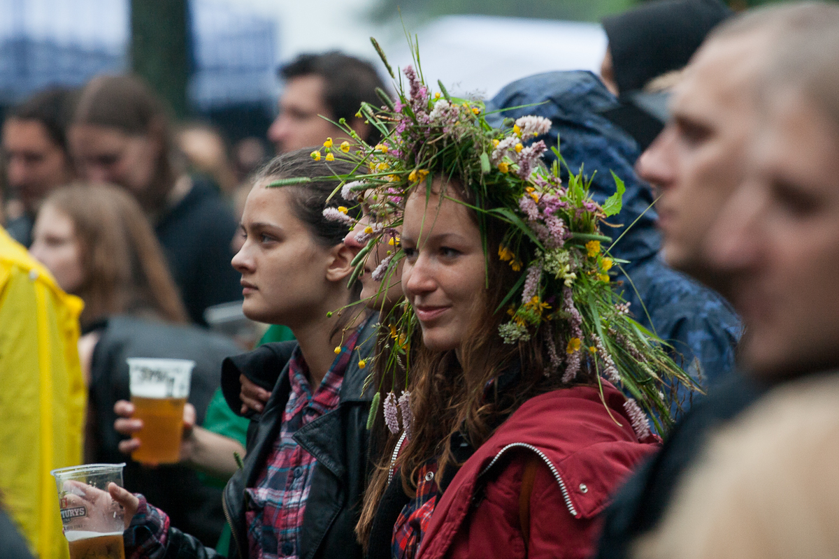 A crowd of people gathered outdoors in 2012, many wearing jackets. In the foreground, a smiling woman wearing a flower crown stands beside another person holding a plastic cup with a beverage. The background shows more individuals at Kilkim Žaibu, some blurred and some focused, indicating various interactions and activities.