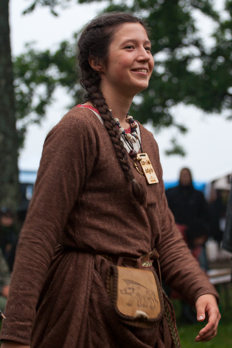 A smiling person with long brown hair in braids, wearing a brown medieval-style dress with a belt pouch and a large necklace. The background features trees, blurred figures, and what appears to be an outdoor festival. The person looks happy and engaged, capturing the festive atmosphere of Kilkim Žaibu 2012.