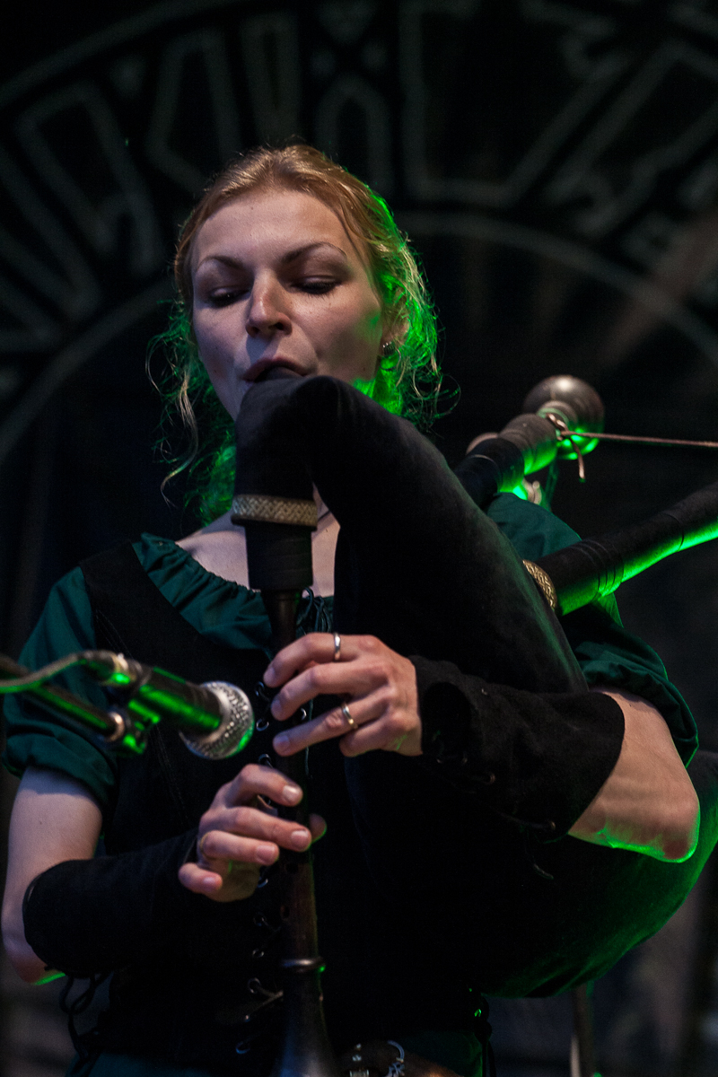 At the Kilkim Žaibu festival in 2012, a woman in a dark green outfit plays the bagpipes with closed eyes, concentrating. The intricate piping is visible. The background features a dark, patterned design illuminated by green light, adding a mysterious ambiance to the scene. She holds the instrument with both hands.