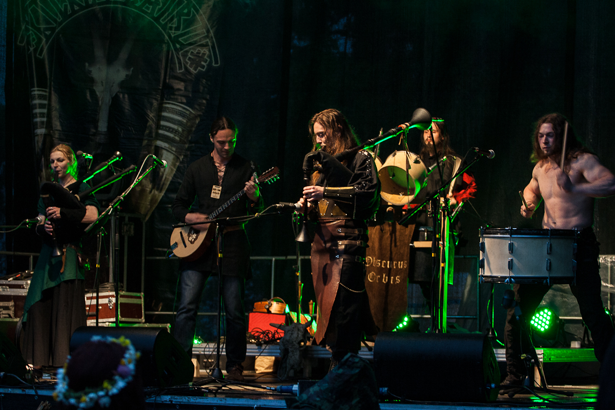 At the 2012 Kilkim Žaibu festival, a band performs on stage with medieval-style instruments. Five members, dressed in medieval-themed costumes, play a variety of instruments including bagpipes, a lute, and drums. The background is dark with green stage lighting and a banner displaying intricate artwork.