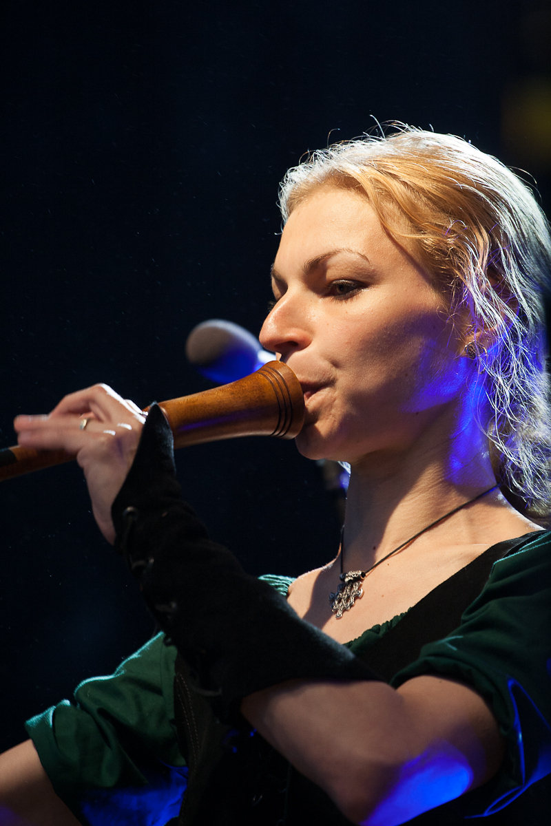 A person with blonde hair is playing a wooden wind instrument on stage at Kilkim Žaibu. They are wearing a green top with long, black fingerless gloves. Blue stage lighting highlights their face, and a microphone is visible in the background, creating an enchanting atmosphere reminiscent of 2012 festival vibes.