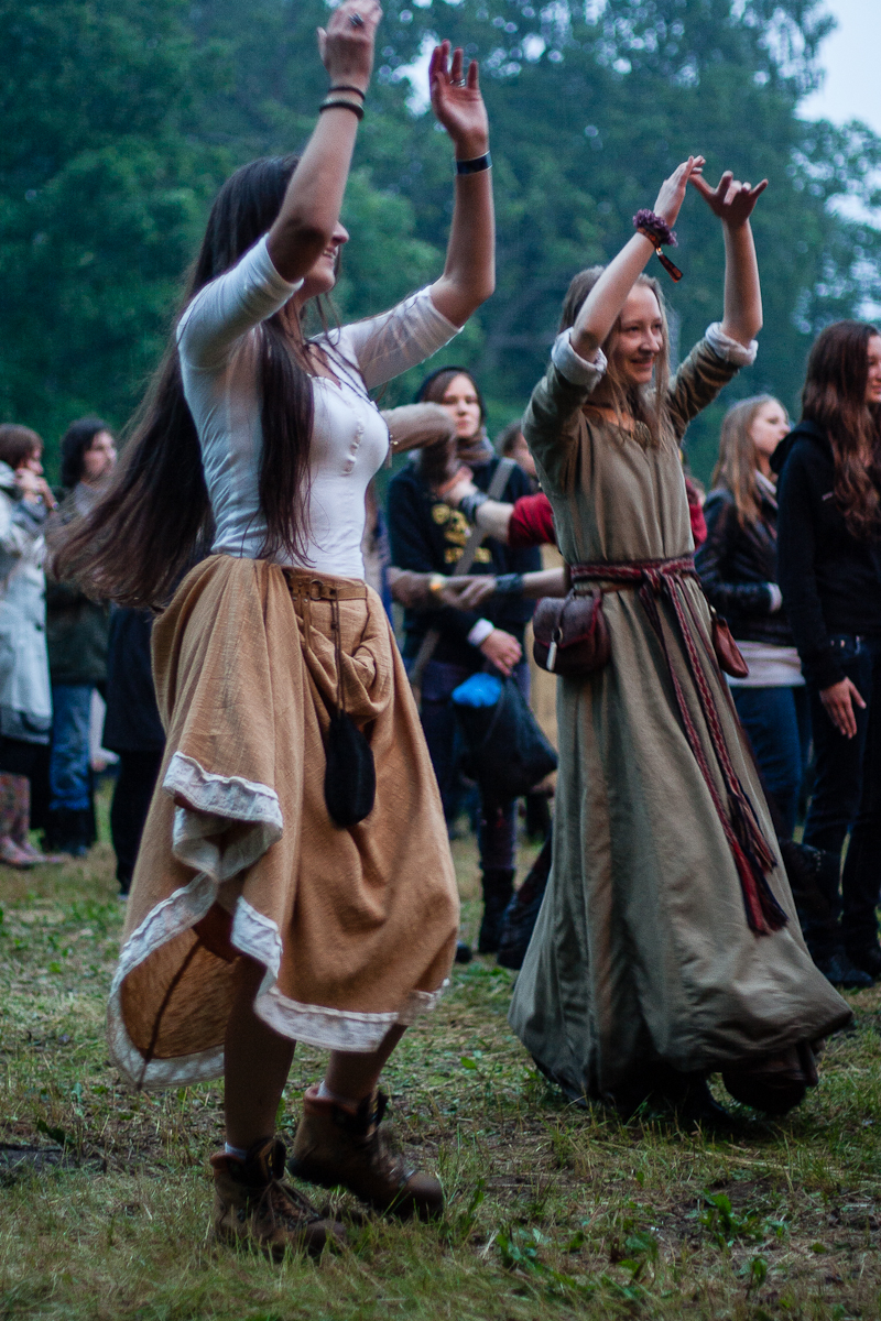 Two women are dancing outdoors at the 2012 festival, dressed in medieval-style clothing. One wears a white blouse with a long beige skirt, and the other wears a green dress with a red belt. They are surrounded by onlookers at what appears to be the Kilkim Žaibu Lithuanian music festival, with trees in the background.