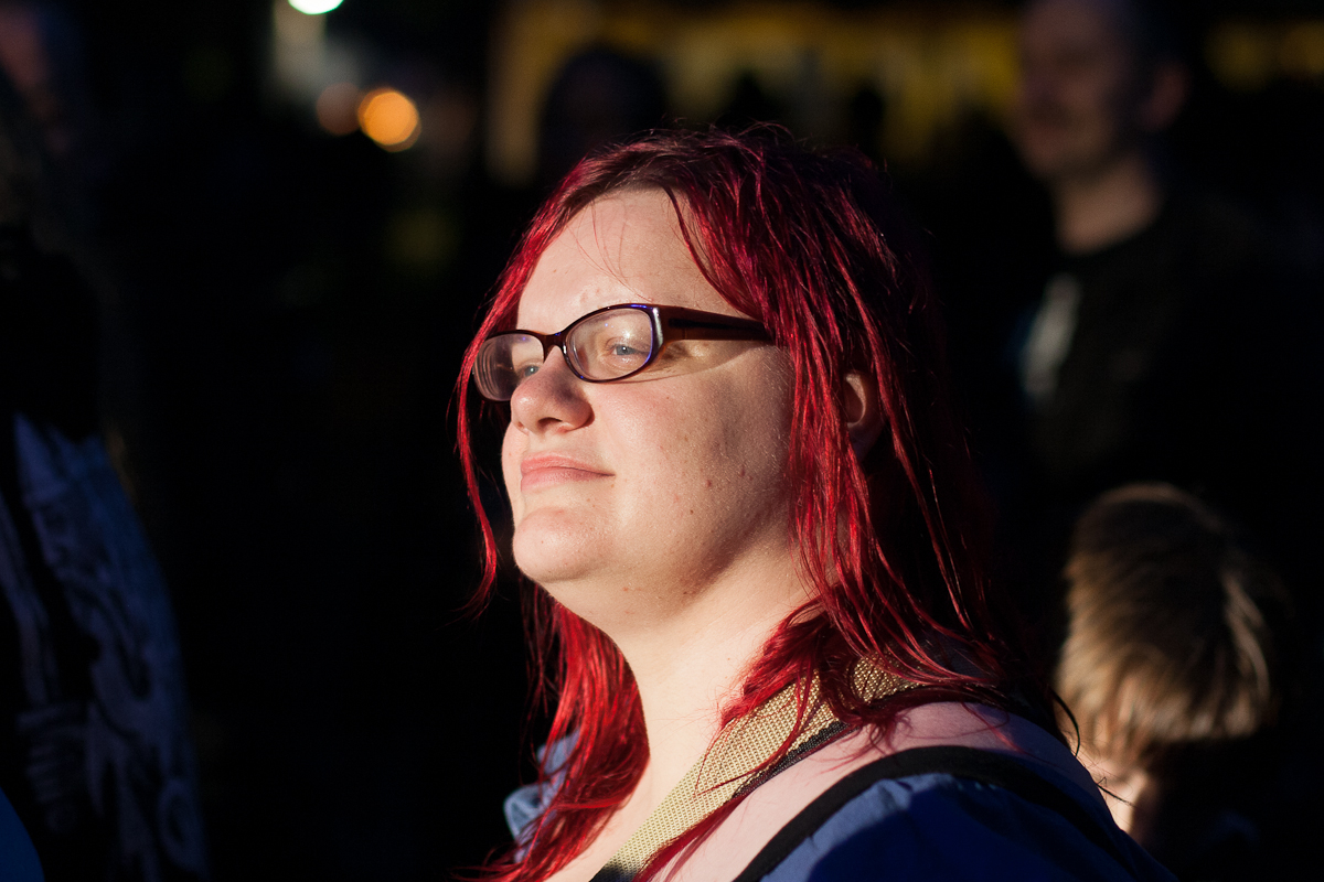 A person with long, bright red hair and glasses stands in a dimly lit area at Kilkim Žaibu 2012, looking to the right with a slight smile. They wear a blue jacket and an over-the-shoulder strap. The background is dark, with blurred lights and indistinct figures, creating a stark contrast with the well-lit subject.