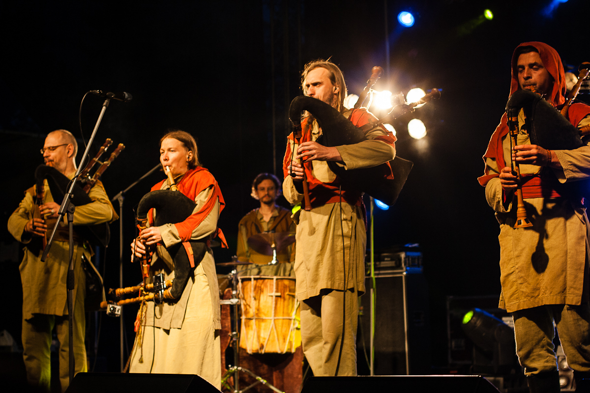 At the 2012 Kilkim Žaibu festival, a folk band performs onstage in medieval attire. Four musicians in beige robes and red hoods play bagpipes, while a drummer in the background plays a large drum. The stage is illuminated by soft yellow lights, with additional colorful lights enhancing the atmosphere.