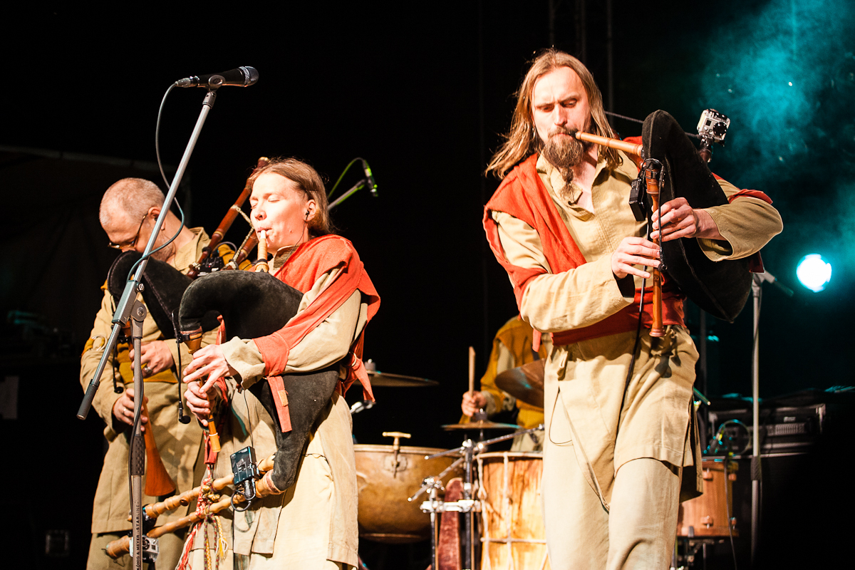 At the 2012 Kilkim Žaibu festival, a musical group performs on stage, featuring three members playing bagpipes and a drummer in the background. All members wear medieval-style costumes in beige and red. They are under stage lighting with a microphone stand visible in the foreground. The background is dark with some green stage lights.