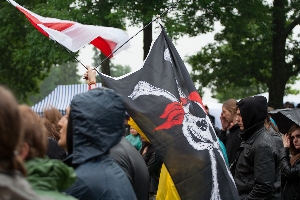 People gather outdoors under cloudy skies at the 2012 Kilkim Žaibu festival. A person in a dark hooded jacket holds a pole with a large black flag featuring a white Jolly Roger with a red bandana, symbolizing a pirate theme. Another flag with red and white stripes is visible in the background, surrounded by green trees.