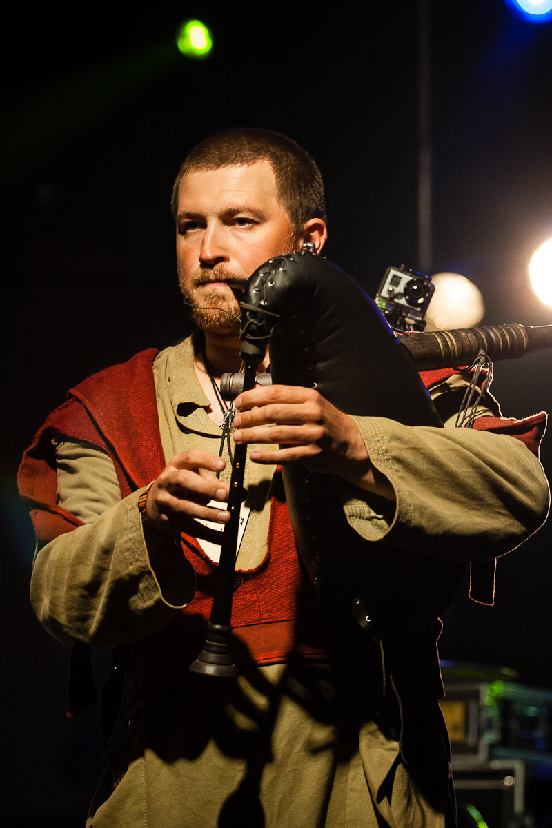 At Kilkim Žaibu 2012, a musician with short hair and a beard is playing a black bagpipe with a wooden pipe on stage. Clad in a medieval-style beige and red tunic, he performs under bright stage lights, which illuminate the dark background. A small camera is attached to his instrument.