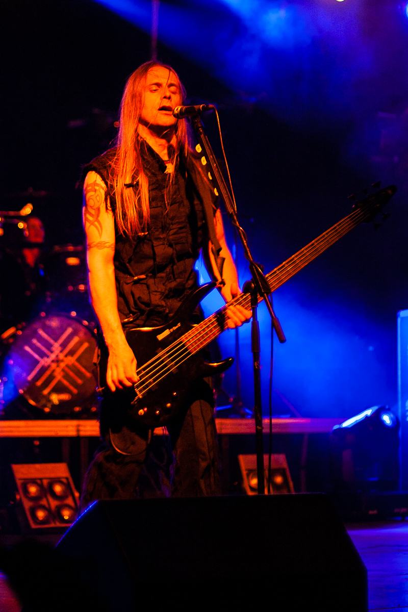 At Kilkim Žaibu 2012, a man with long hair performs on stage, playing a black electric bass guitar and singing into a microphone. He wears a sleeveless black top, revealing tattoos on his arms. Blue and purple stage lights create a dynamic atmosphere, while the drum kit with its logo is blurred in the background.
