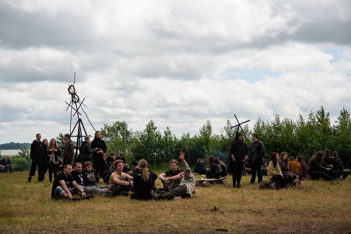 A group of people dressed in medieval attire sit and stand on a grassy field under a cloudy sky during Kilkim Žaibu 2012. Some engage in conversation, while others gaze around. Wooden structures with wheel-like designs stand in the background, and a treeline is visible further back.