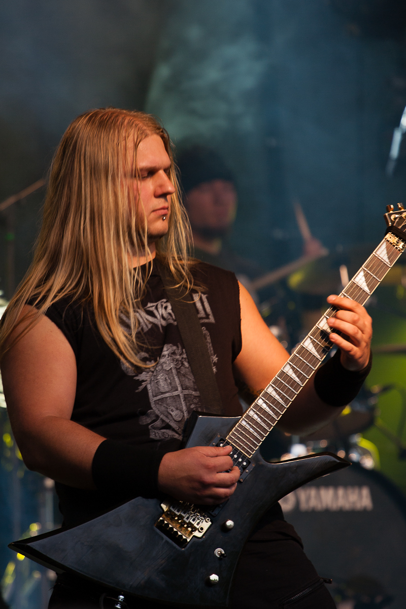 A musician with long blonde hair, wearing a black sleeveless shirt with a graphic design, plays an electric guitar on stage at the 2012 Kilkim Žaibu festival. He has a serious expression, and the background features a drummer, drums, and a "Yamaha" bass drum. The stage is dimly lit with blue and green lighting effects.