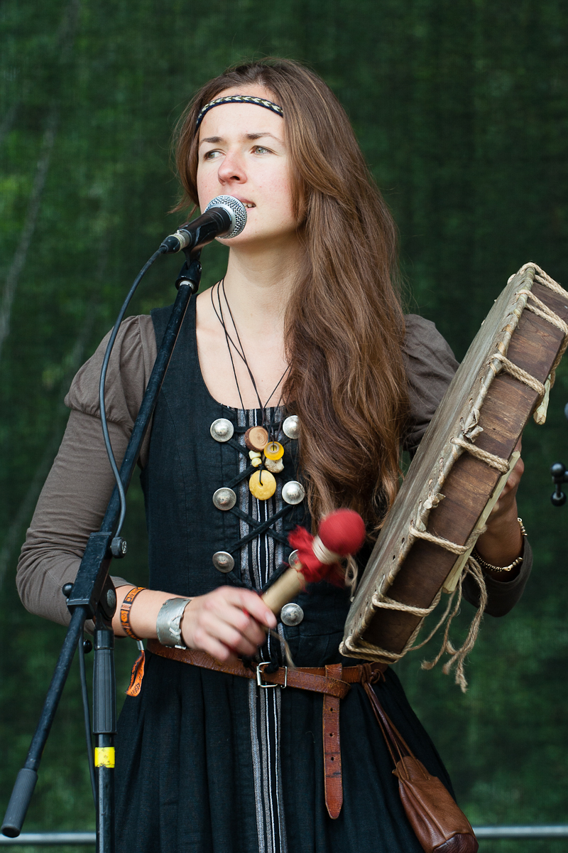 A woman with long brown hair stands on stage at the Kilkim Žaibu Festival, wearing a black dress with silver buttons and a brown shirt underneath. She is holding a handheld drum in her left hand and a mallet in her right, singing into a microphone in front of a green background. She wears a narrow headband and several necklaces.