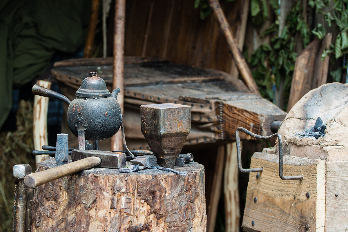 A blacksmith's workspace featuring a weathered anvil, various hammers, and tools scattered on a tree stump. Nearby is a teapot with a handle and spout. The background displays wooden structures and green foliage, reminiscent of the rustic ambiance at Kilkim Žaibu 2012.