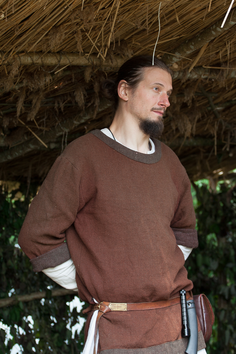 A person with long dark hair tied back and a light beard is standing under a thatched roof at the 2012 Kilkim Žaibu festival. They are wearing a brown tunic over a white shirt and have a belt with a leather pouch and a sheathed knife at their waist. They are looking to the right with their hands clasped behind their back.