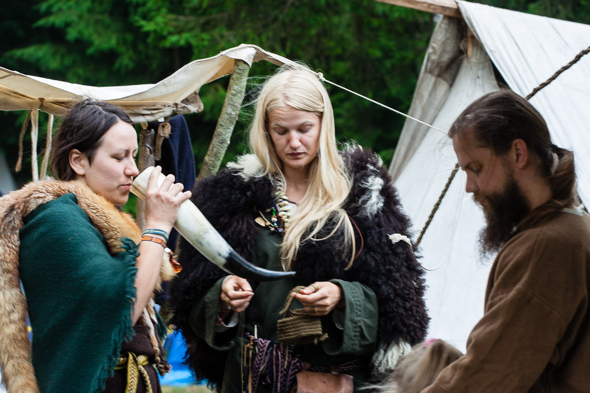 Three people are dressed in medieval or Viking-style clothing in front of a canvas tent at Kilkim Žaibu 2012. The person on the left drinks from a large horn, the one in the middle looks down while holding an object, and the person on the right has their head bowed, perhaps focusing on something. Trees are visible in the background.