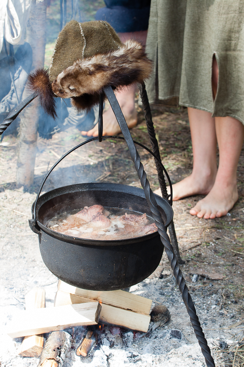 A black cast iron pot hangs over an open fire, suspended by a tripod made of twisted metal rods. The pot contains simmering food. A fur-lined hat rests on top of the tripod. Barefoot individuals wearing historical garments stand on the grass in the background, capturing a moment reminiscent of Kilkim Žaibu 2012.