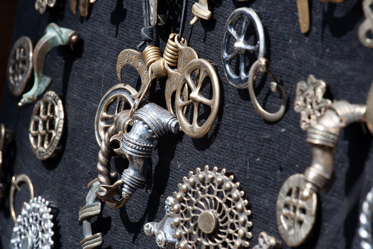 Close-up image of various steampunk-style metal trinkets and gears attached to a black textured background. The items include cogs, keys, wheels, and intricate mechanical parts, showcasing a blend of industrial and Victorian aesthetics reminiscent of the Kilkim Žaibu 2012 festival. The metallic pieces vary in color, primarily bronze and silver.