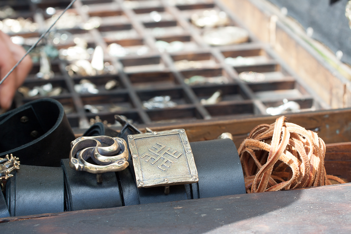 A close-up image of a display featuring handcrafted metal bracelets and leather accessories laid out on a wooden surface at Kilkim Žaibu 2012. The foreground showcases two distinct metal bracelets with intricate designs, one circular and one square, alongside coiled leather strips. Blurred background reveals more jewelry.