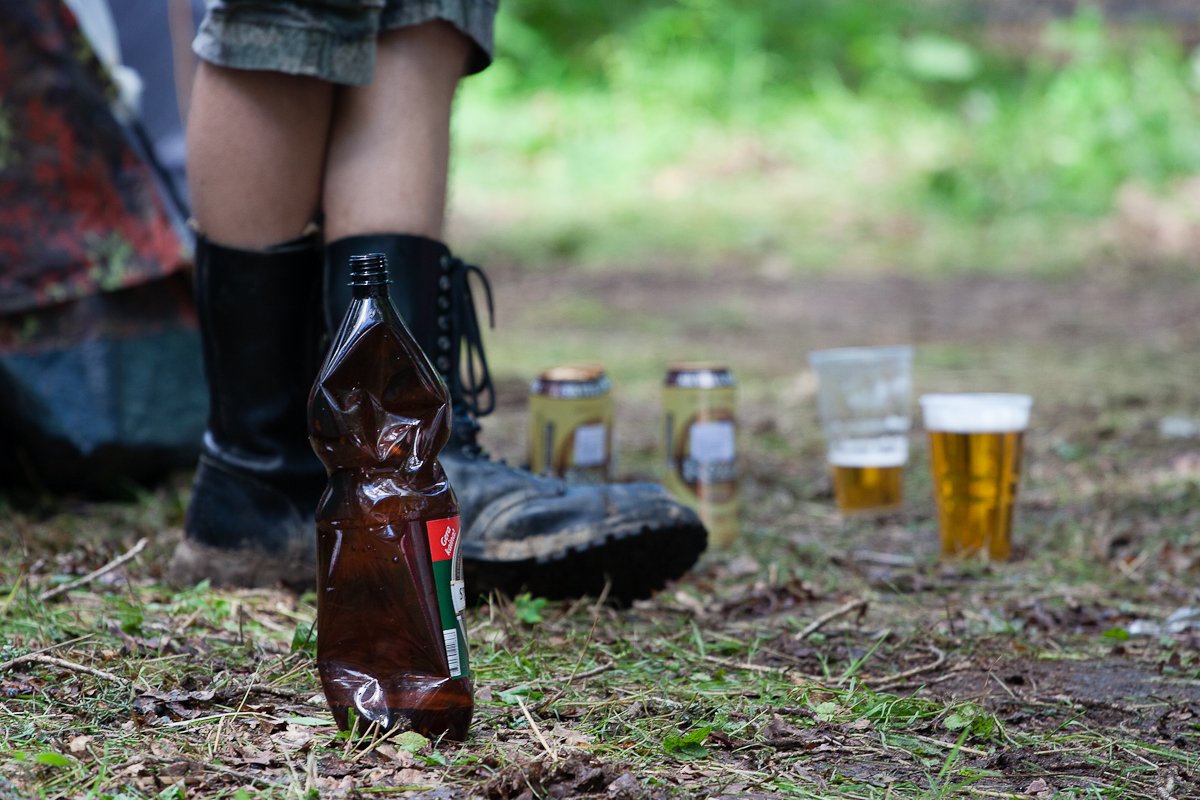 Close-up of a person wearing black rain boots and shorts standing on a forest floor with scattered leaves and grass at the 2012 Kilkim Žaibu festival. In the foreground, a crumpled brown plastic bottle lies on the ground. In the background, several beer cans and plastic cups filled with beer are scattered about.