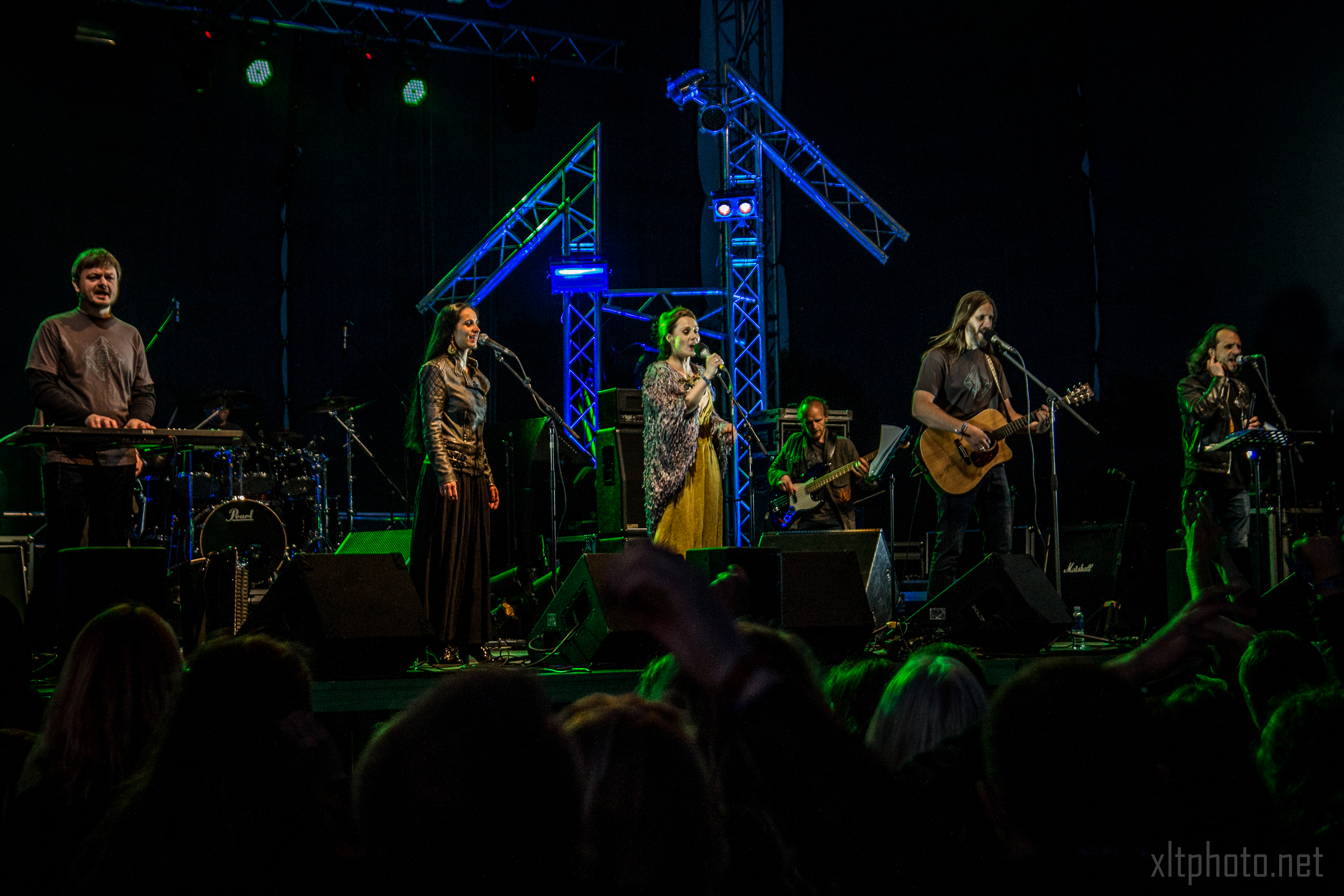 A band performs on a dark stage featuring colorful lights and rigging at the 2015 Kilkim Žaibu festival. The group comprises five members: three women, one man with a guitar, and another man with a keyboard. Audience members can be seen in the foreground, some with raised hands. The backdrop includes various musical equipment.