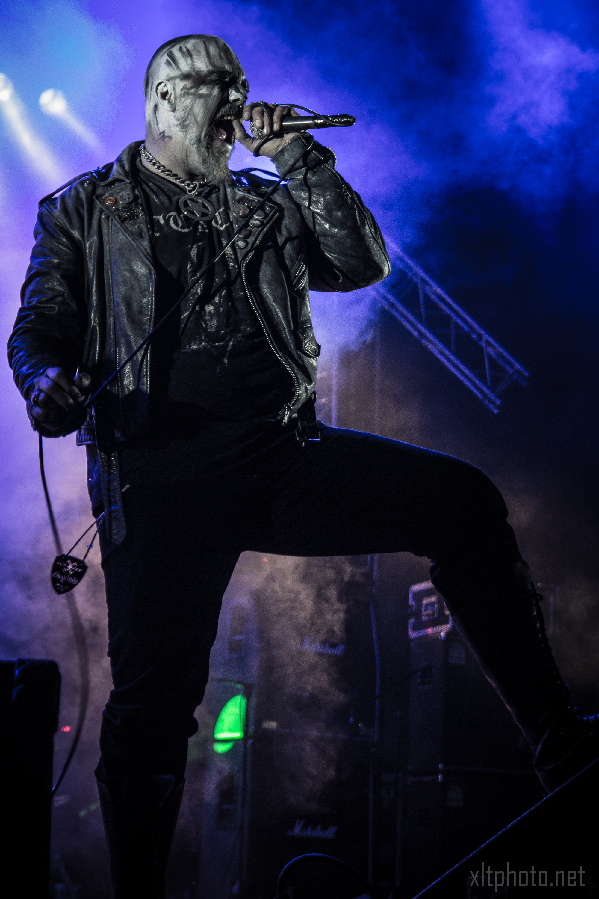 A person in a black leather outfit and dramatic face paint passionately sings into a microphone on a dimly lit stage at the Kilkim Žaibu festival. Blue and purple lights illuminate the background, musical equipment including speakers and guitars are visible, and smoke adds a mysterious atmosphere, enhancing the performance mood.