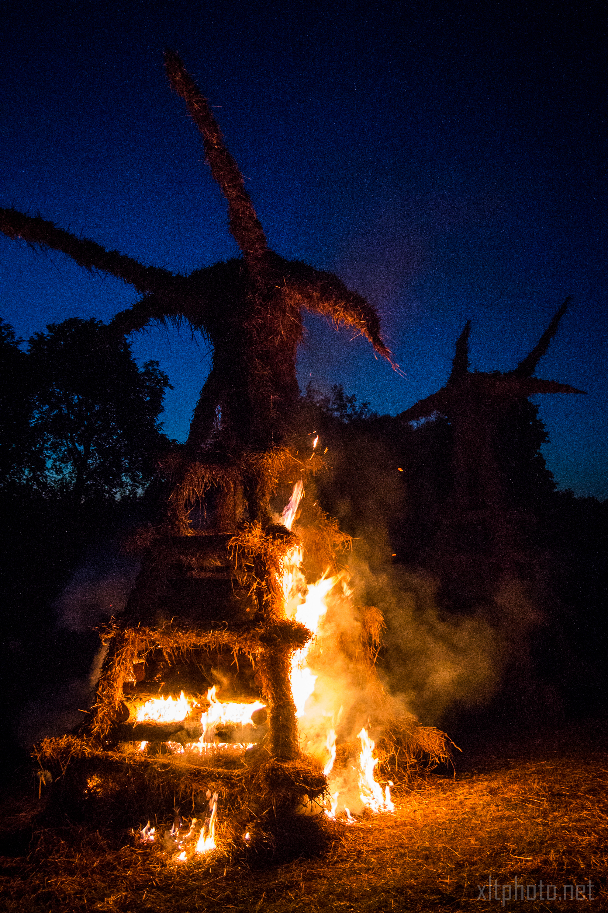At the Kilkim Žaibu festival in 2015, a tall, human-like sculpture made of straw is engulfed in flames at night, set against a dark blue sky. The burning straw emits bright orange and yellow flames, creating a stark contrast with the surrounding darkness. Another similar but unlit sculpture is visible in the background.
